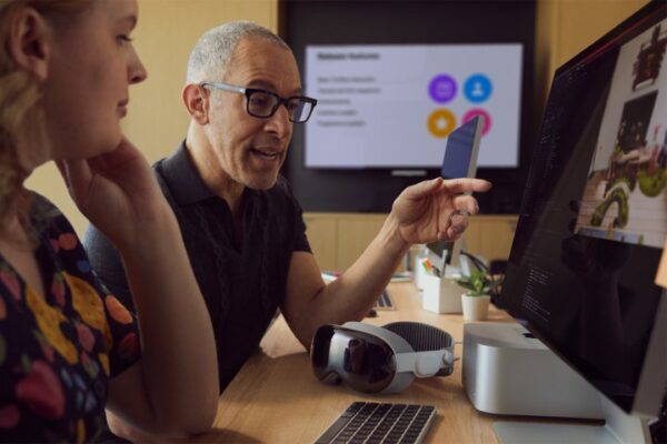 A developer points to a Mac screen while a Vision Pro rests on the desk. A developer points to a Mac screen while a Vision Pro rests on the desk.