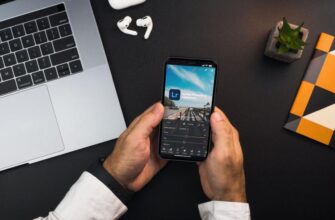 Мужчина держит смартфон с приложением для редактирования фотографий от Adobe на фоне ноутбука, наушников и блокнота.