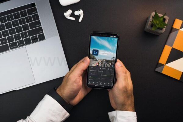 Мужчина держит смартфон с приложением для редактирования фотографий от Adobe на фоне ноутбука, наушников и блокнота.