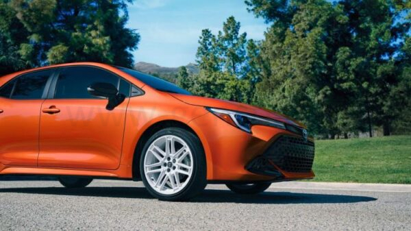 Статическая фотография передней части оранжевой Toyota Corolla Hatchback FX Edition 2026 года на фоне деревьев.