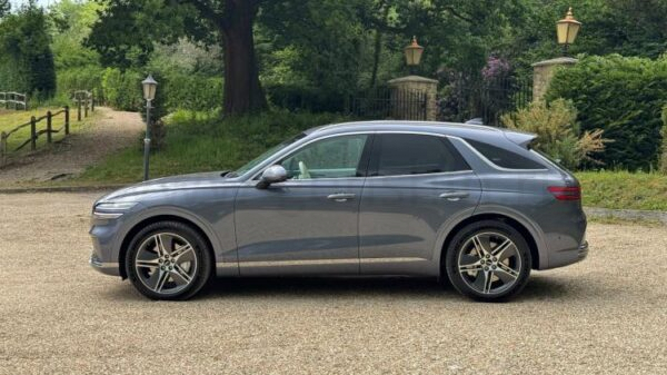 Static side profile shot of a gray 2026 Genesis Electrified GV70 parked on gravel with trees in the background.