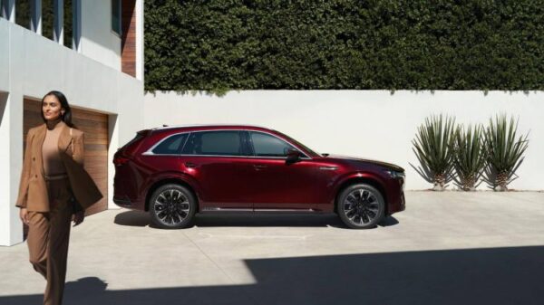 Static side profile shot of a red 2026 Mazda CX-90 parked on a drive with a woman walking in the foreground.