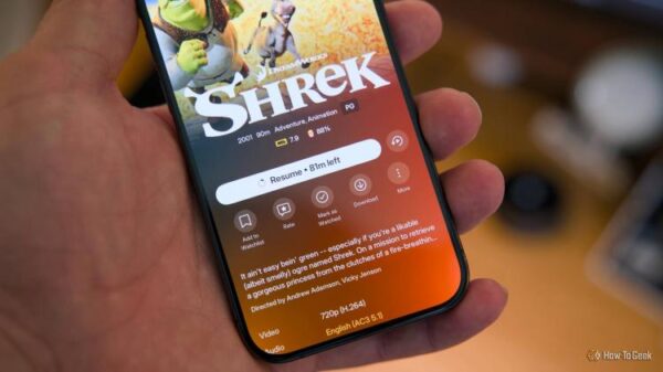 В руке держат iPhone 15 Pro с открытым Plex и значком скачивания на фильме Шрек.