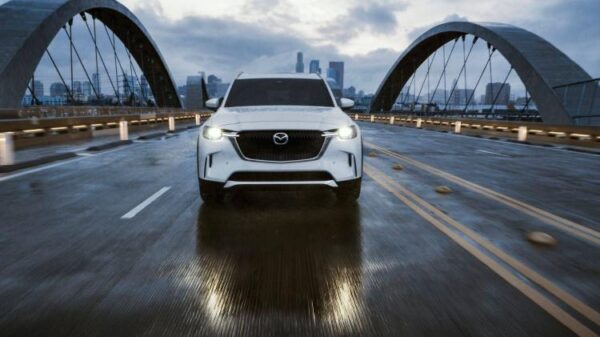 Динамичный вид спереди на белую Mazda CX-90 2026 года, движущуюся по мосту на фоне города.