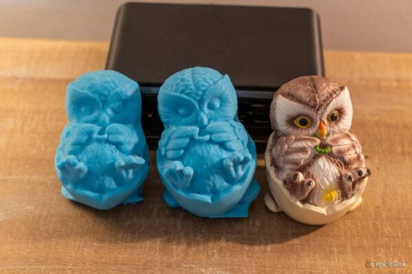 Two blue 3D-printed owl scans beside the original painted owl figurine with an iPhone behind them.